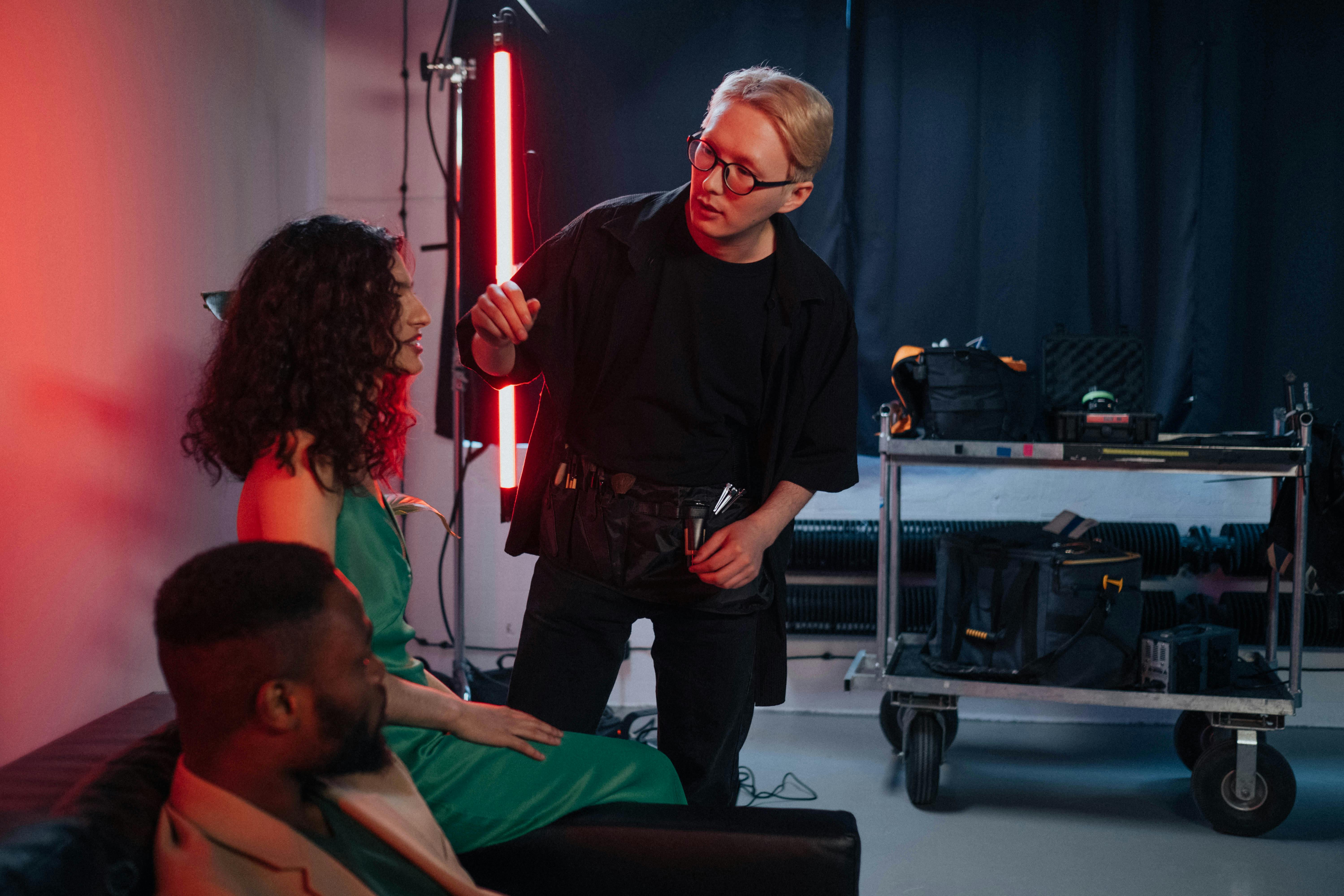 Film crew filming an actor on a couch in a red-lit studio with cameras, a clapperboard, and production lights.