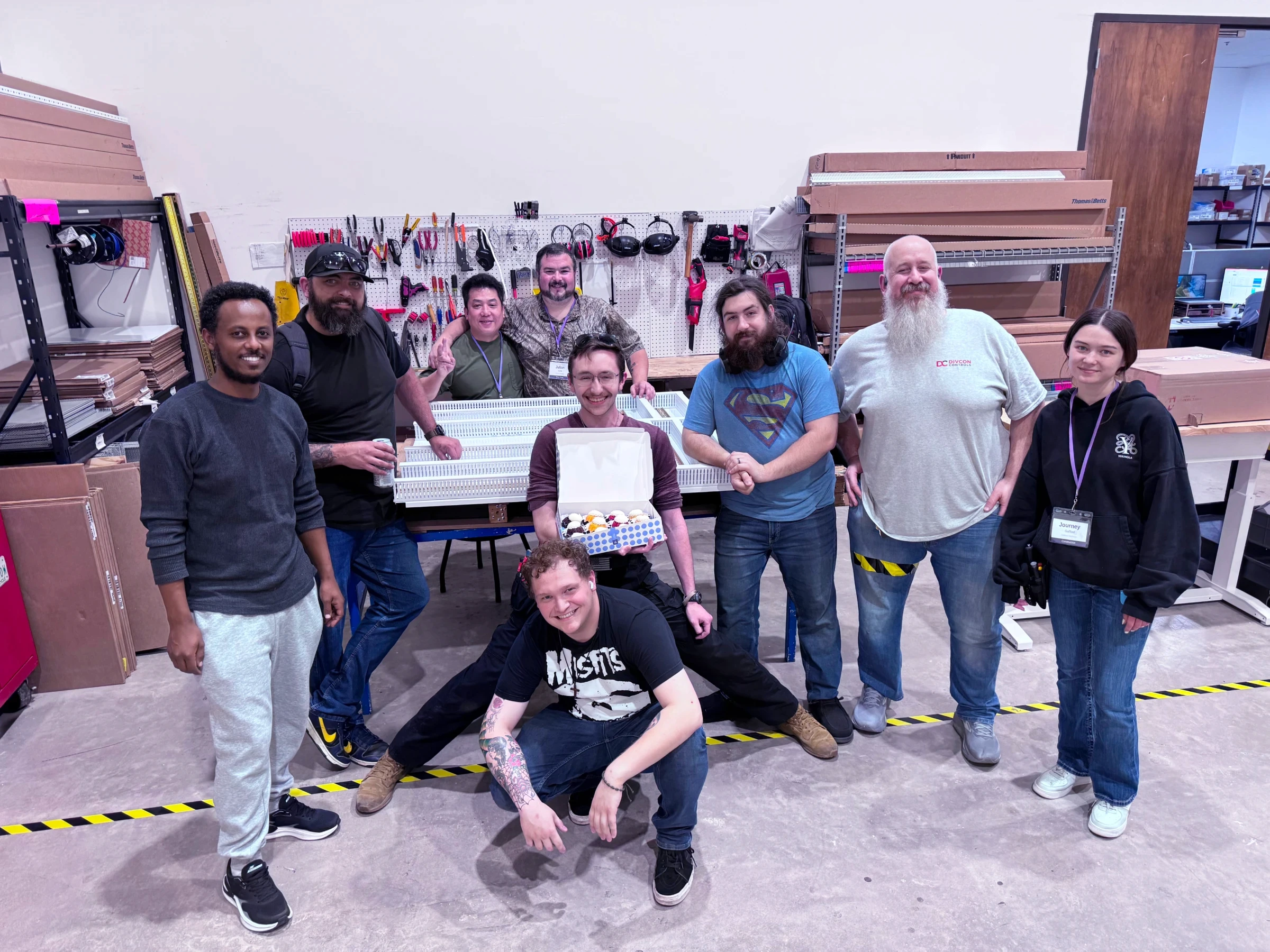 Nine coworkers pose together in a workshop, with one holding an open box of treats.