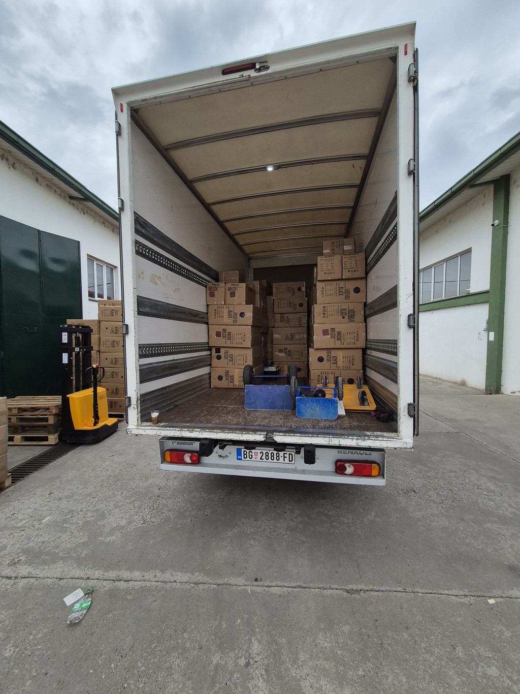 Kamion za transport robe parkiran u magacinu, utovaren kartonskim kutijama složenim do plafona i opremljen ručnim paletarom i kolicima za prevoz.