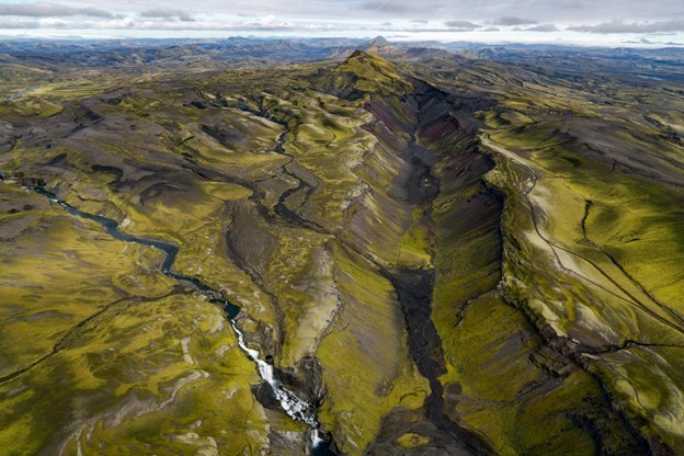 the picture shows the Eldgjá canyon in Katla Geopark, in the Highlands of south Iceland from the air. The Eldgjá volcanic fissure eruption that formed this canyon was the largest on earth in the past 1000 years, and the canyon itself stretches 40 kilometers in length. There is a long, lush green canyon, and black and red lava beds and sands. A waterfall pours down the side of the mossy canyon.