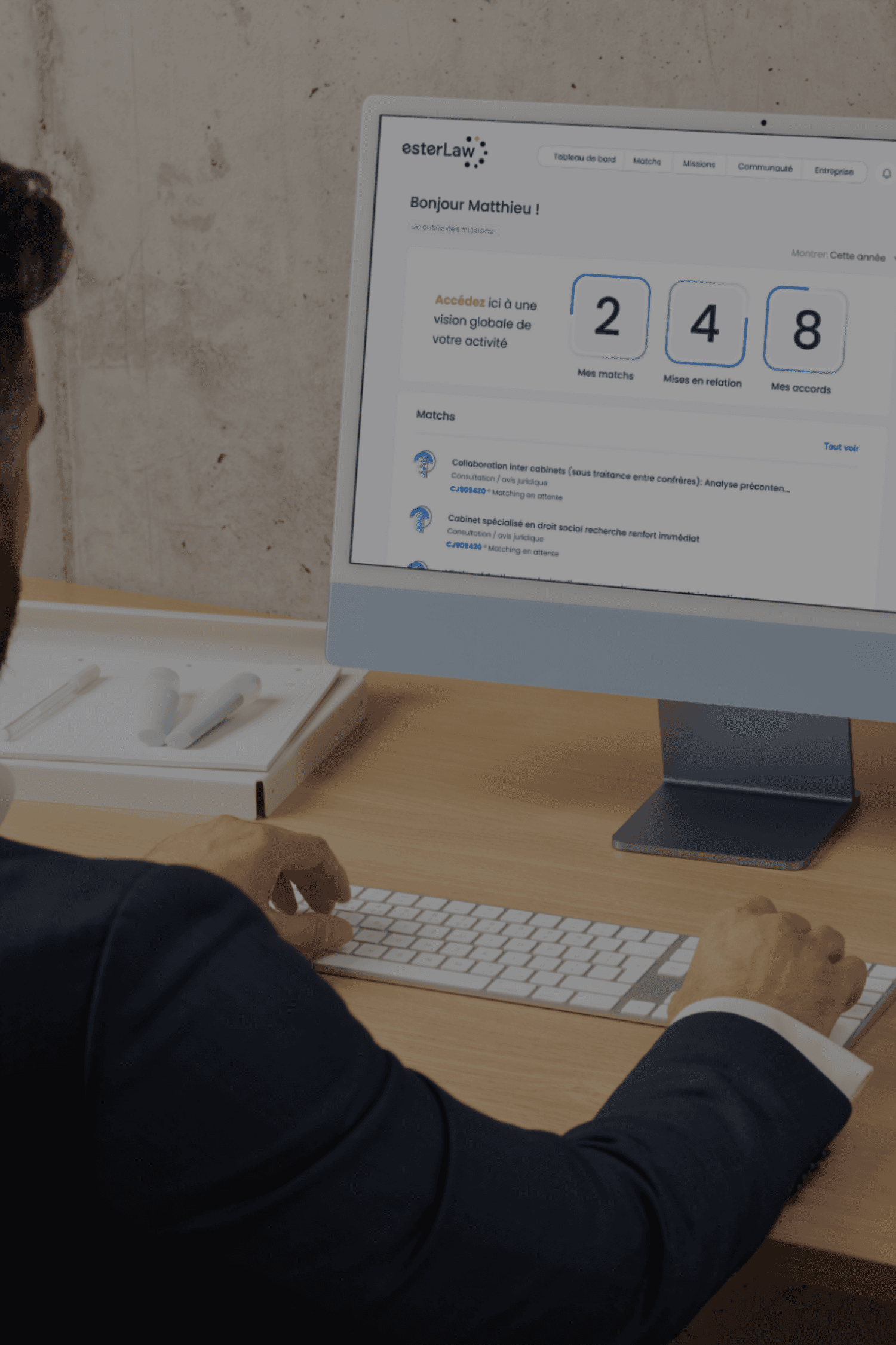 A man in a grey shirt sits in a dim office, gazing seriously at a computer screen. Blinds and office supplies create a focused and professional atmosphere.