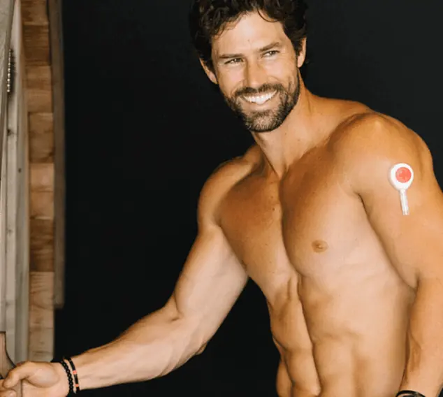 Shirtless man smiling indoors, showing a wearable health patch on his upper arm.