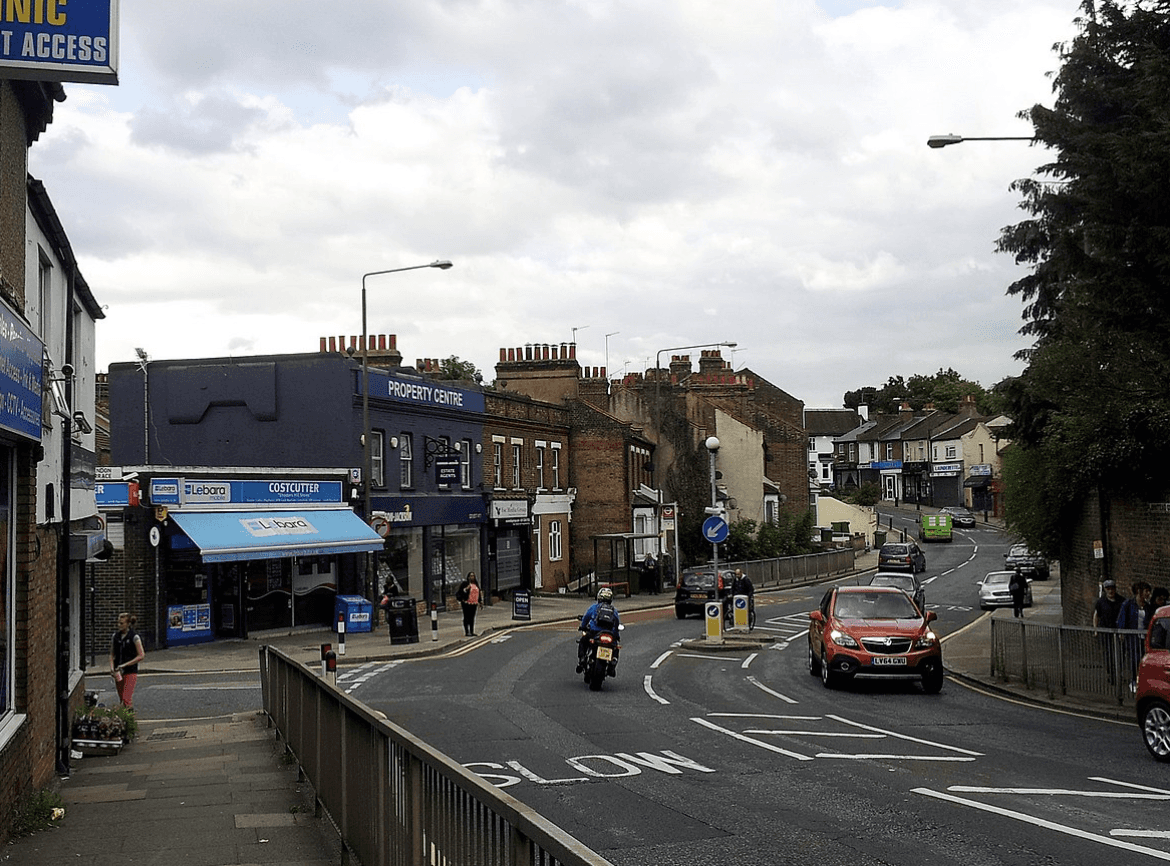 A smiling man with curly hair is sitting at a desk, looking at the camera, with a laptop and a lamp in the background.