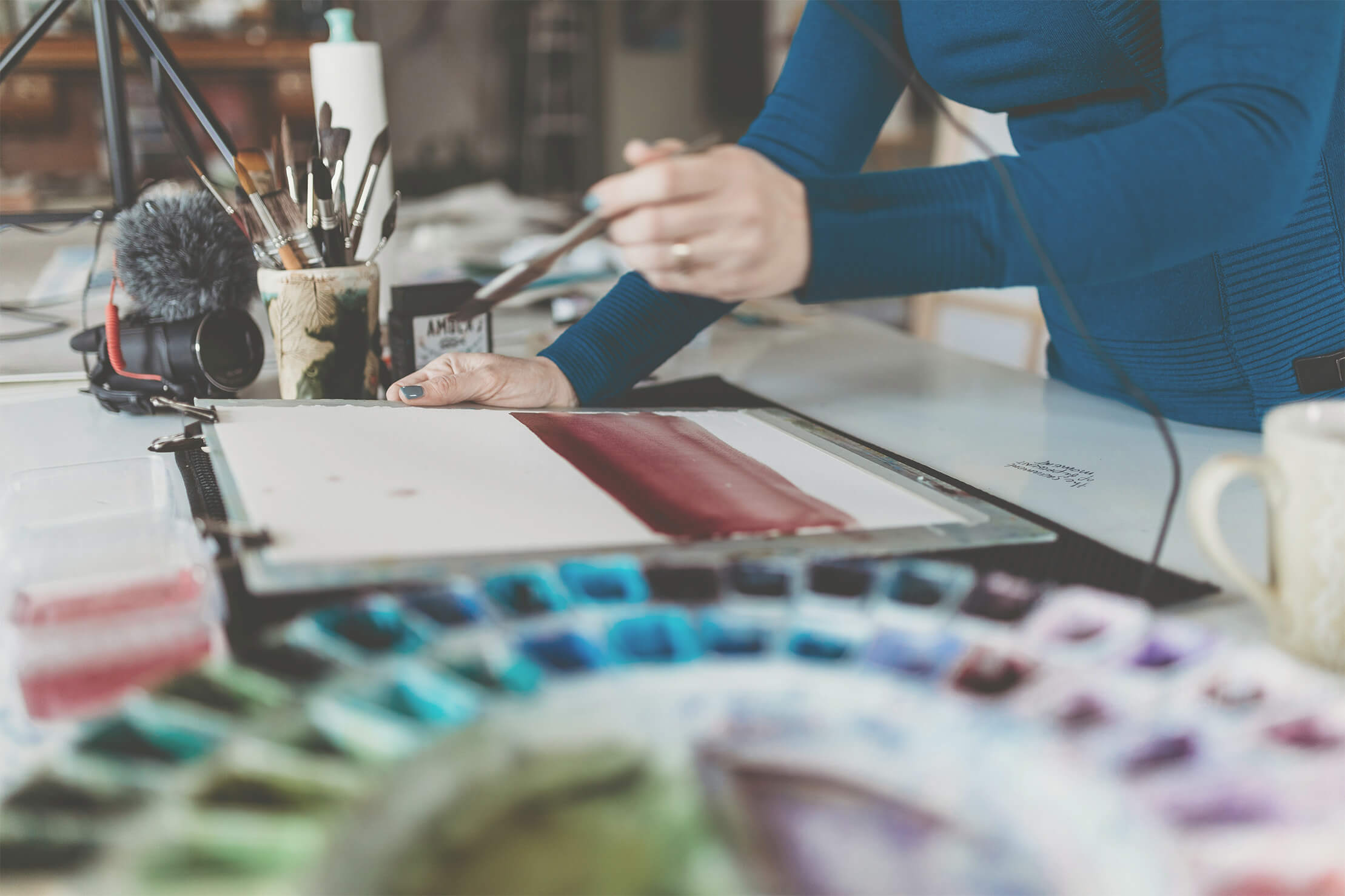 Artist working on watercolor painting with paint brushes and palette on desk.
