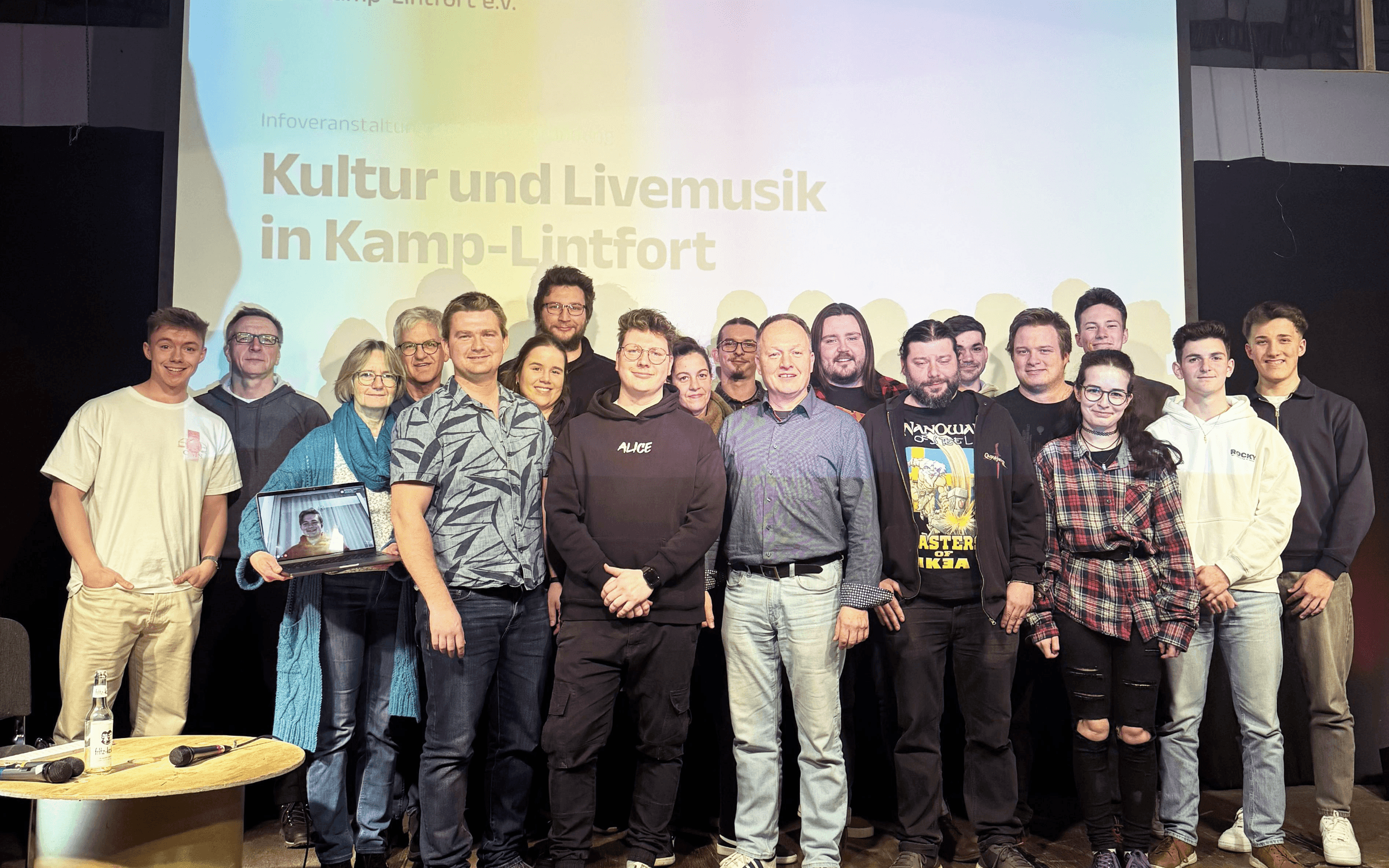 „Mehrere Personen des KlangRevier-Teams stehen gemeinsam auf einer Bühne vor einer großen Leinwand mit der Aufschrift ‚Kultur und Livemusik in Kamp-Lintfort‘. Die Gruppe wirkt freundlich und präsentiert sich als Teil des Festivals.