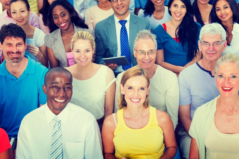 A diverse group of real people of various ages smiling together in a bright, crowded setting. The image emphasizes authentic human connection and a sense of belonging, highlighting the "human touch" that defines a brand community in an increasingly digital and AI-driven world.