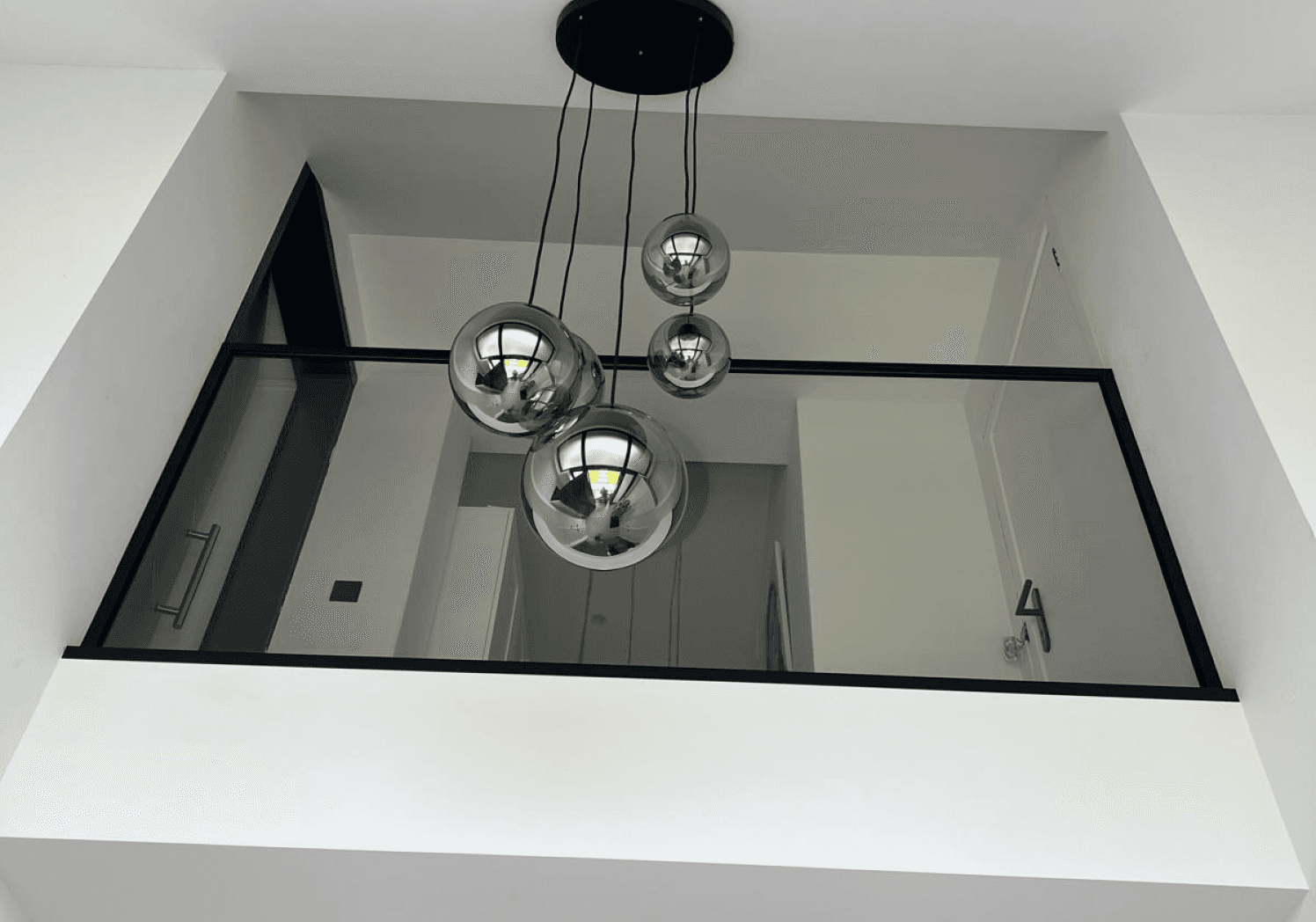 Opwaartse blik op een modern interieur, mogelijk een trappenhuis of atrium, met een cluster metalen bolvormige hanglampen aan het plafond en een glazen balustrade rond een opening naar de lagere verdieping, wat een gevoel van hoogte en openheid geeft.