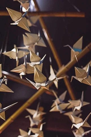 Dekoracja z zawieszonych papierowych żurawi origami, podświetlonych ciepłym światłem i zawieszonych na drewnianej konstrukcji