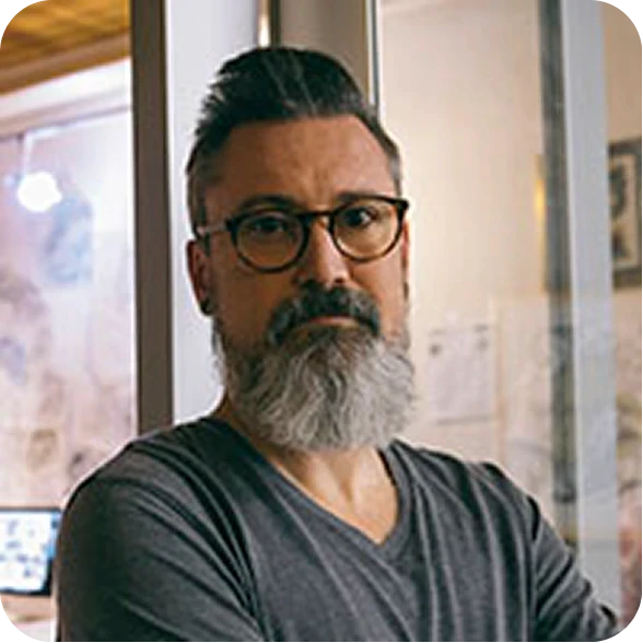 Serious mature man with a graying beard and glasses looking directly forward in a brightly lit indoor office space.