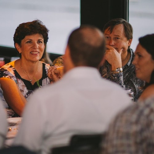 Apat na tao ang nakaupo sa paligid ng mesa habang nag-uusap, tatlo ang nakikita, kabilang ang isang babaeng nakangiti at dalawang lalaki.