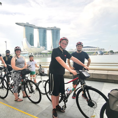 กลุ่มนักปั่นจักรยานโพสท่าพร้อมกับจักรยานริมฝั่งน้ำ โดยมี Marina Bay Sands เป็นฉากหลัง ภายใต้ท้องฟ้าส่วนที่มีเมฆบางส่วน