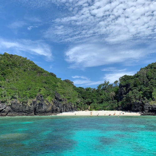 A pristine beach with lush green hills, clear turquoise water, and a few people relaxing under a partly cloudy sky.