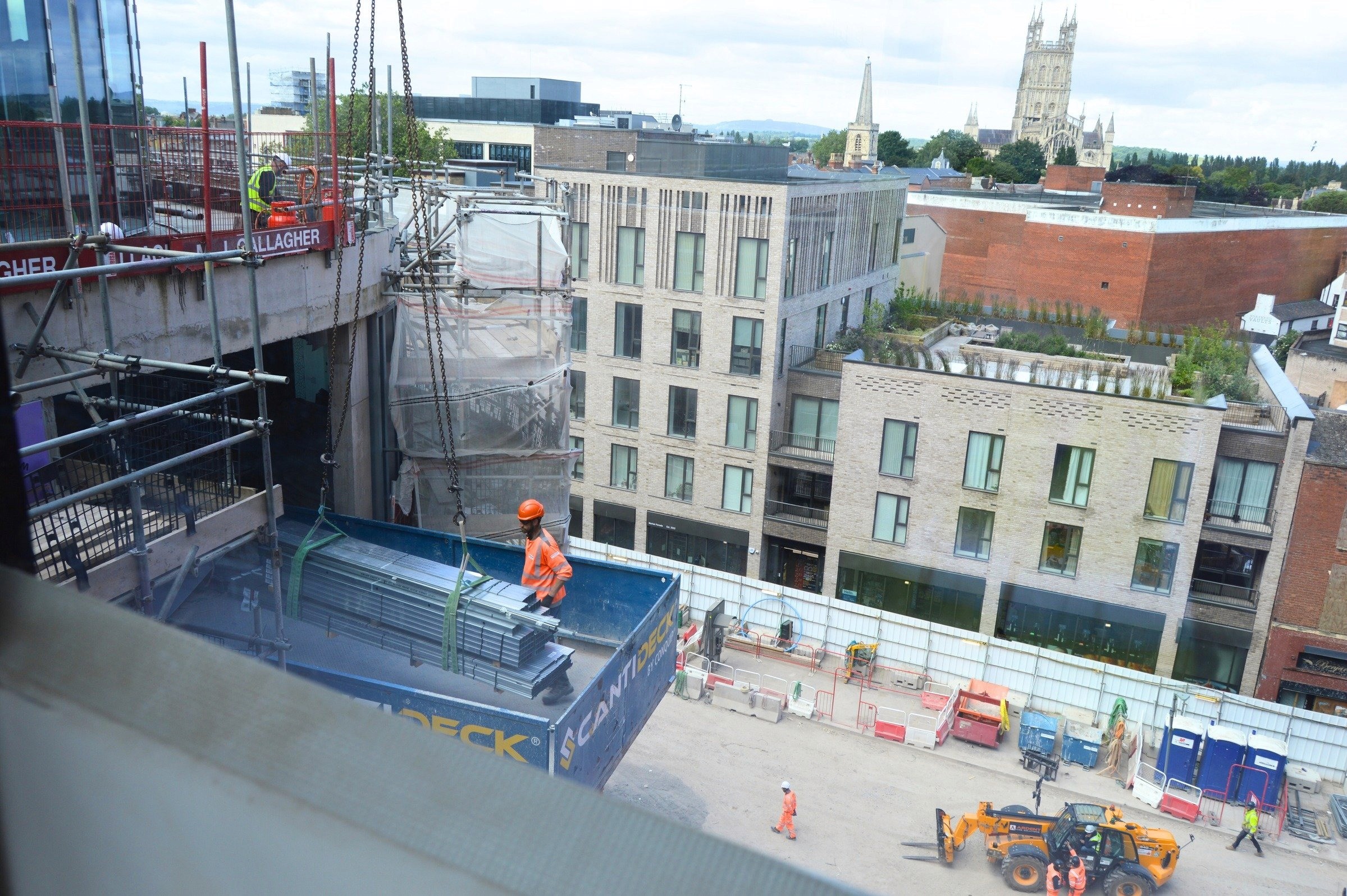 A construction site with workers and equipment, featuring a large crane lifting metal beams into a multi-story building, with an urban cityscape and a historic cathedral visible in the background.