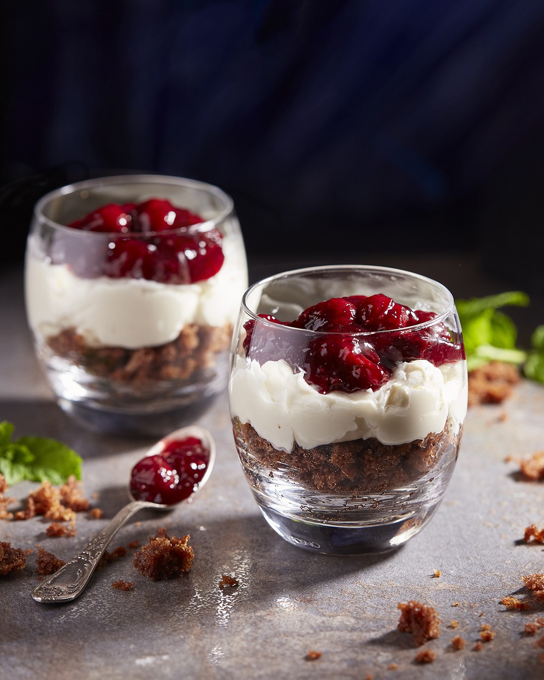 Two glass cups filled with layers of chocolate crumbs, creamy white yogurt, and a topping of vibrant red cherry compote are artfully arranged on a textured gray surface, with a spoon and scattered crumbs enhancing the elegant dessert presentation.