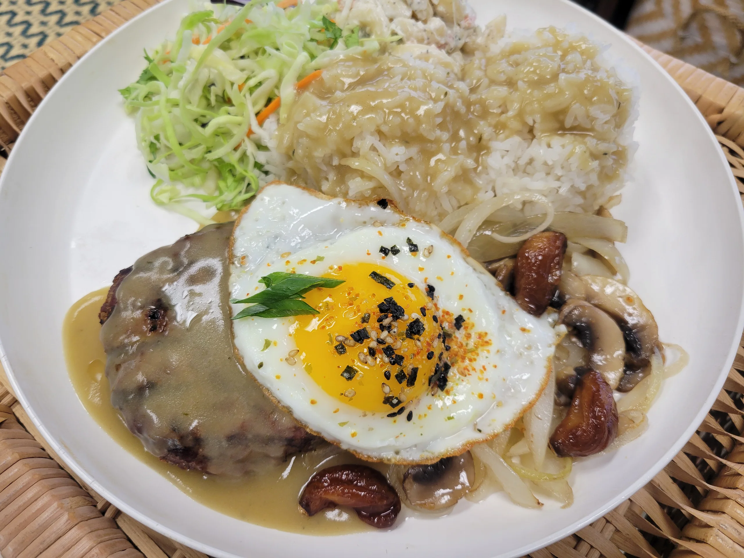 A char-grilled Angus beef patty topped with a sunny-side-up egg and thick mushroom gravy, served with rice and mac salad in a bowl.
