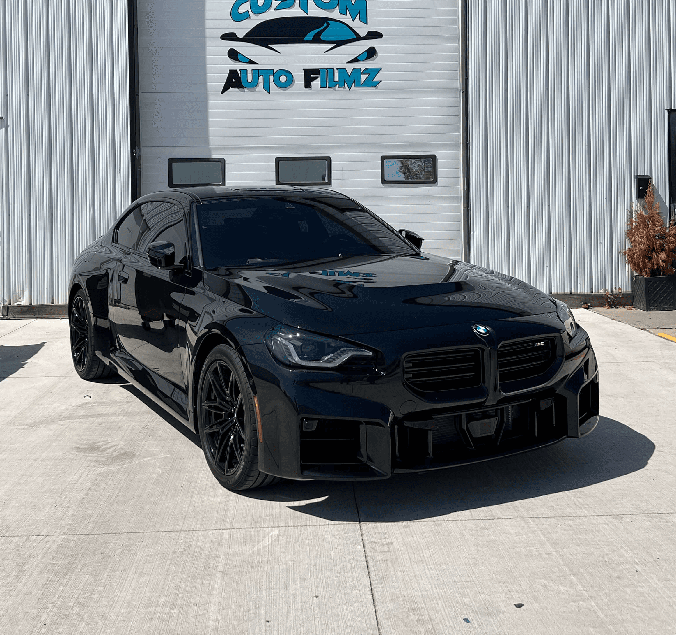 A black BMW M2 sports car is parked on a concrete driveway in front of a white industrial building.