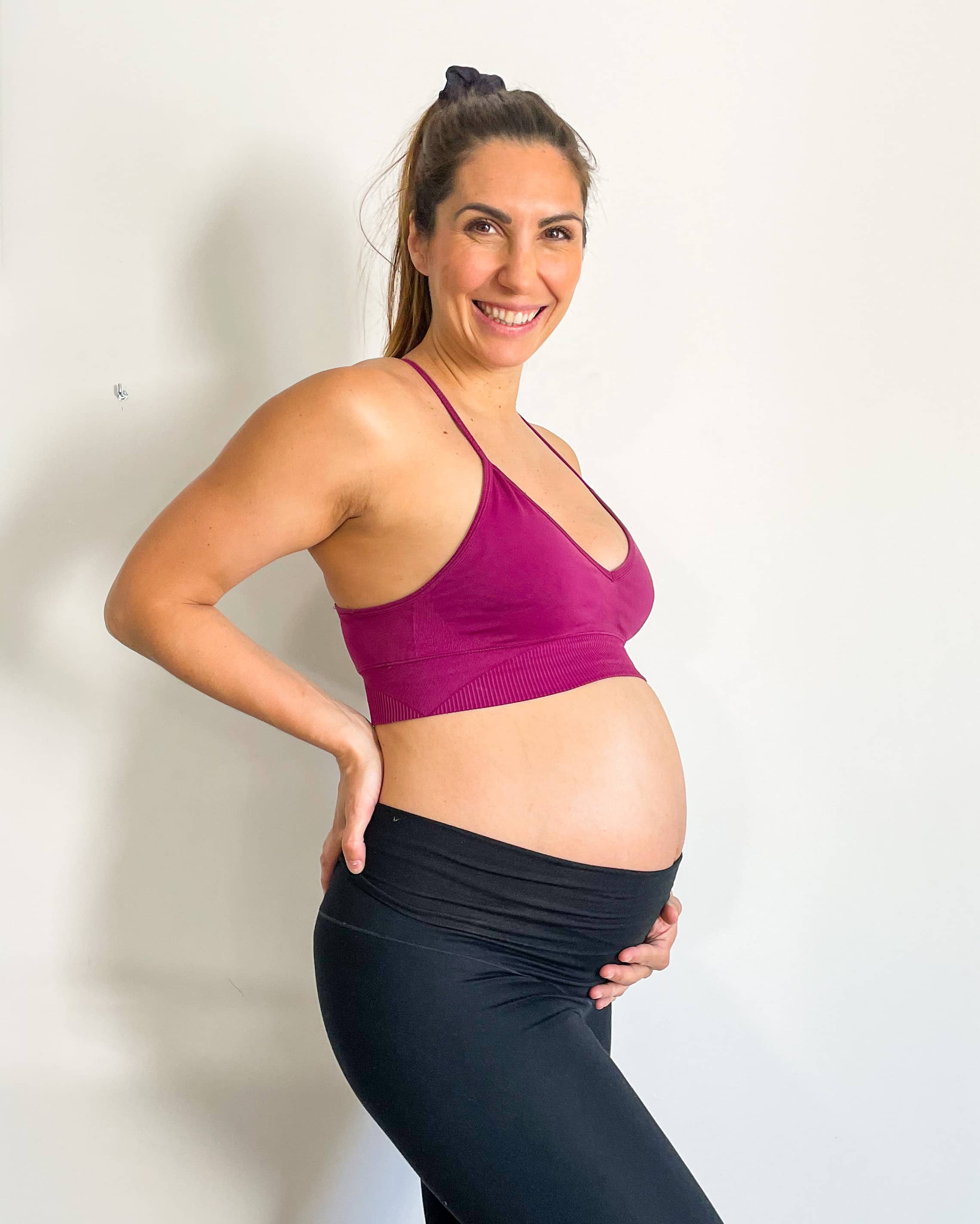 Portrait studio de Caroline Rinaudo, femme enceinte souriante avec mains sur son ventre et le dos, posant en tenue de sport noire et violette sur fond blanc.