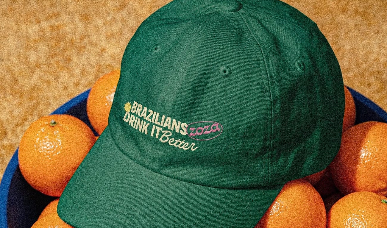 Boné verde escuro com os dizeres "Brazilians drink it better" em amarelo claro. O boné está em cima de laranjas dentro de um balde azul.