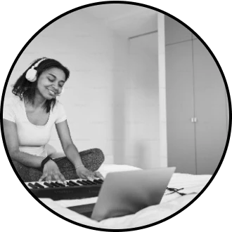 A keyboardist is jamming online, playing a synthesizer, using a laptop and headphones, and sitting on a bed.