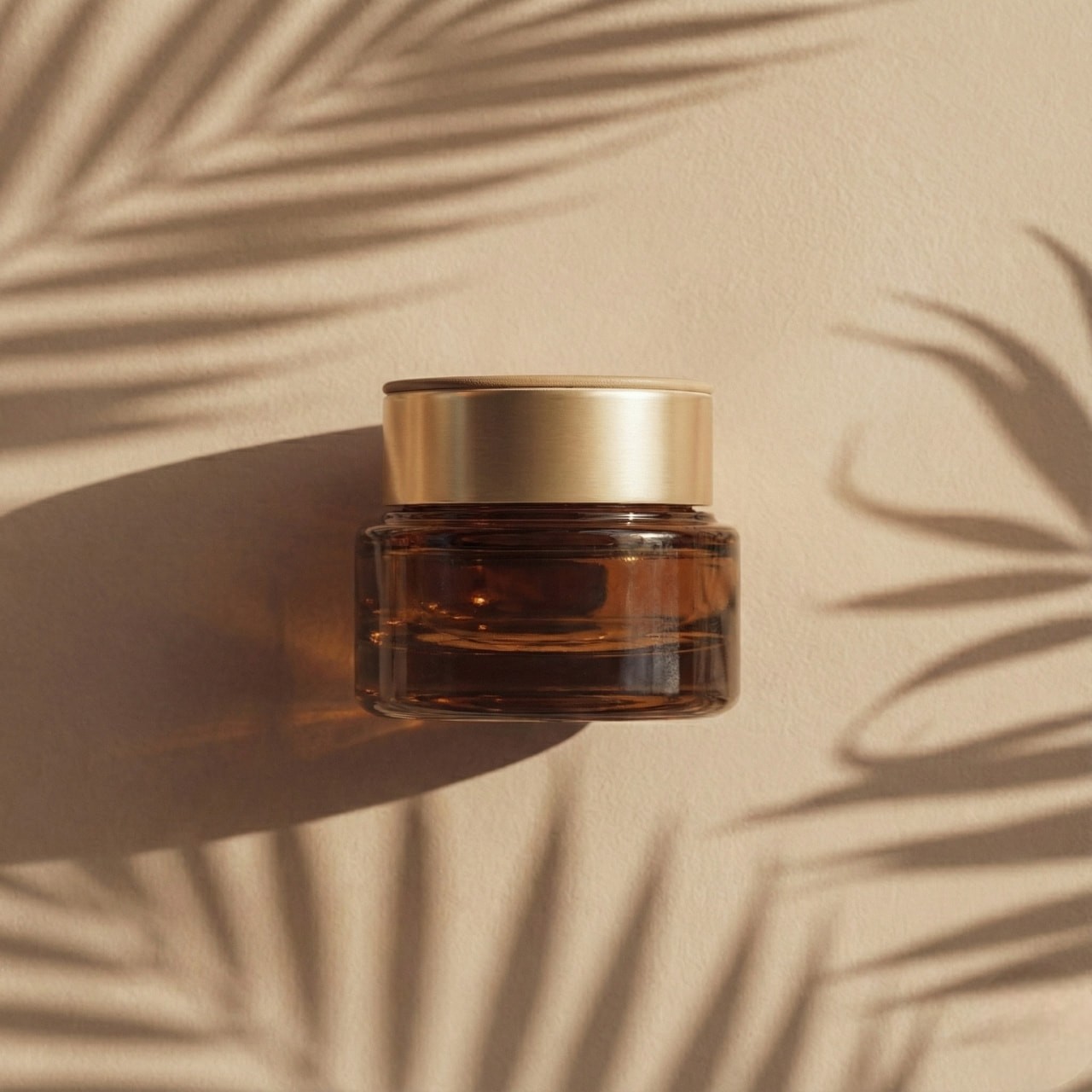 An amber glass jar with a gold lid casts a shadow on a beige surface. Palm leaf shadows surround it, creating a warm, tropical vibe.