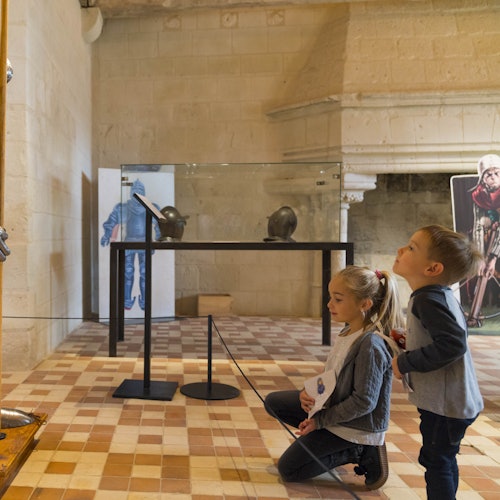 Dos niños en un museo observan cascos medievales expuestos en una vitrina. La sala tiene paredes de piedra y un suelo de baldosas.