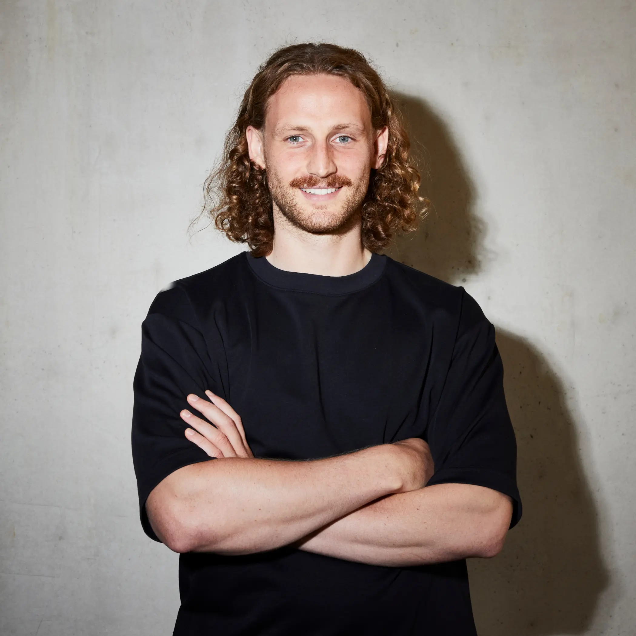 Ein Mann (Robin Junglas) mit langem, lockigem Haar und Schnurrbart steht vor einer kahlen Wand. Er trägt ein schwarzes T-Shirt, hat die Arme verschränkt und lächelt.