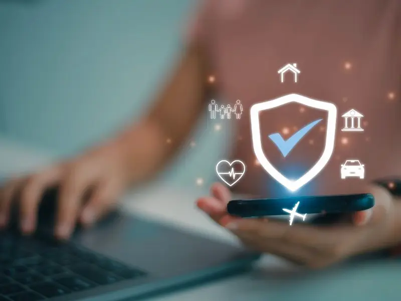 In the image, we see a close-up of a person using a laptop and holding a smartphone. Above the phone, a holographic image of a shield with a checkmark is displayed, surrounded by icons representing different aspects of insurance and protection, such as family, health, home, finance, vehicle, and travel. The visual effect suggests a futuristic or advanced technology concept for comprehensive personal security or insurance coverage.