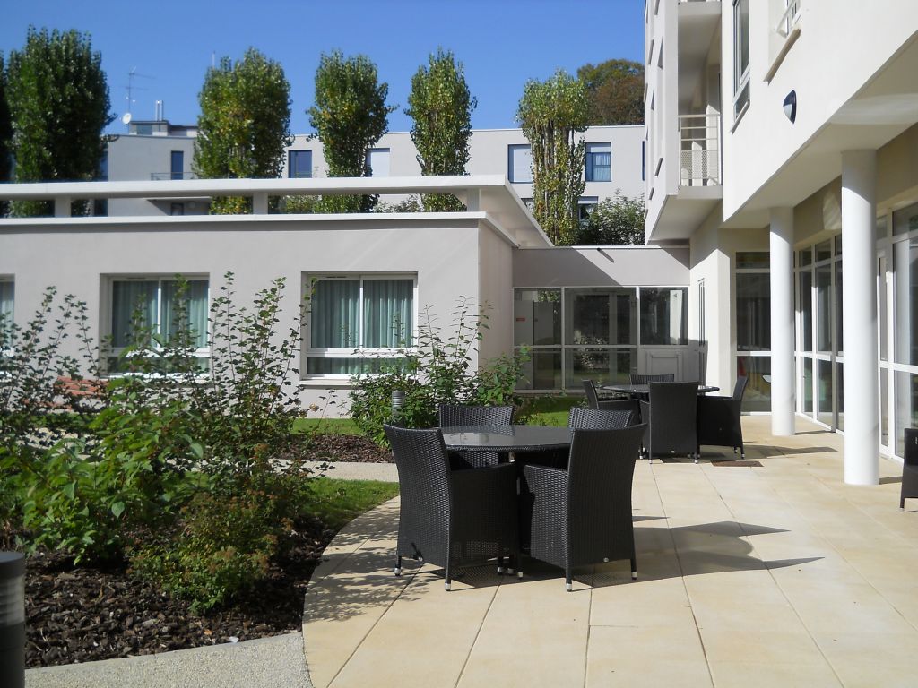 Modern white residential building entrance with green vegetation, paved courtyard area, and blue sky