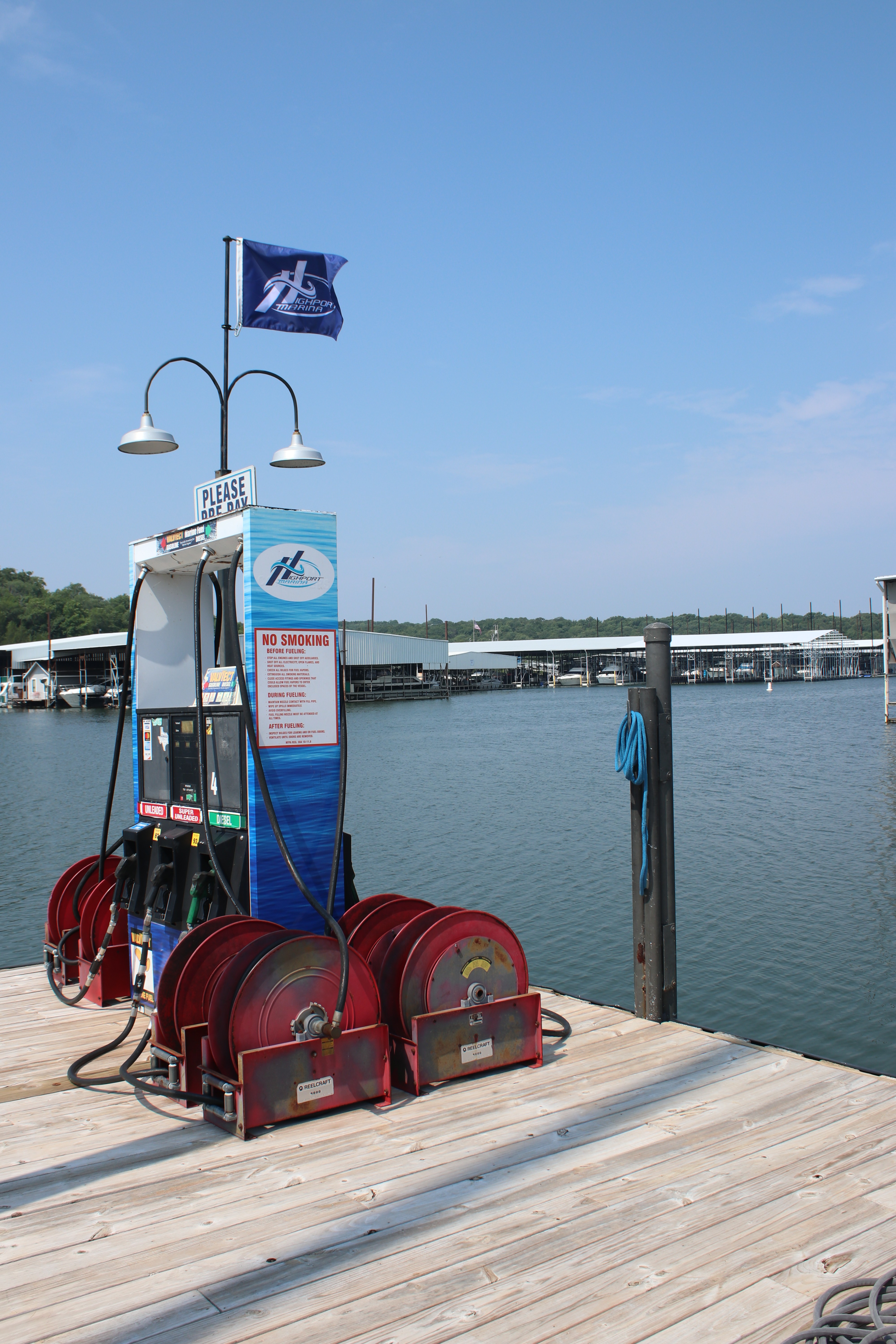 A marina fuel station with multiple fuel hoses is positioned on a wooden dock, surrounded by calm water and boat docks in the background under a clear blue sky.