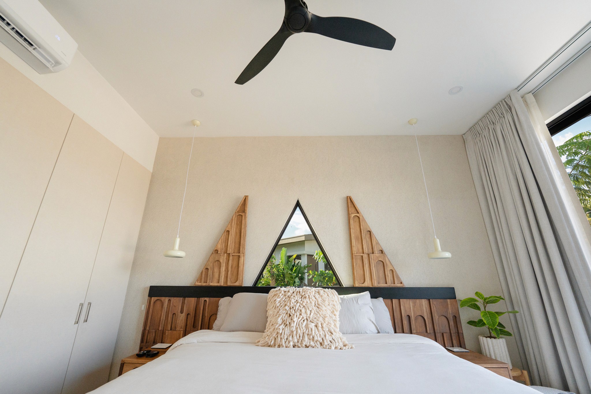 Detail view of a luxury casita bedroom featuring a minimalist design, neutral tones, and unique geometric wood wall art. The room includes bedside pendant lighting, air conditioning, and a view of the lush Costa Rican landscape.