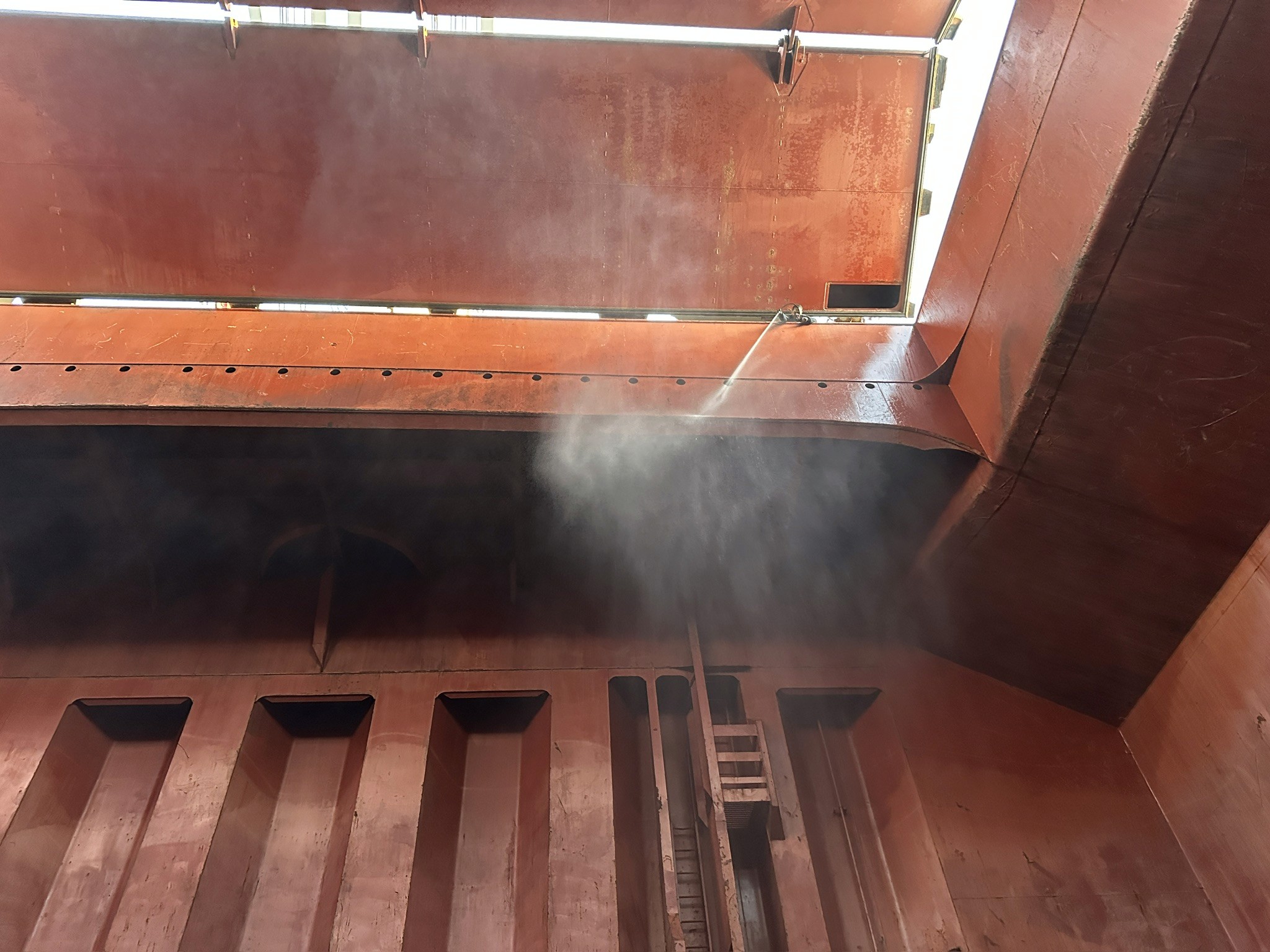 Professional high-pressure washdown in progress inside a large cargo hold. Spray nozzle directed at the corrugated tanktop surface with water mist and droplets visible. Steel structural frames, longitudinal members, and bilge well drains are visible in the foreground. The hold shows orange-red primer coating on upper surfaces and darker weathered steel on lower areas, demonstrating typical pre-cleaning vessel condition. Image exemplifies the active cleaning phase of professional cargo hold preparation.