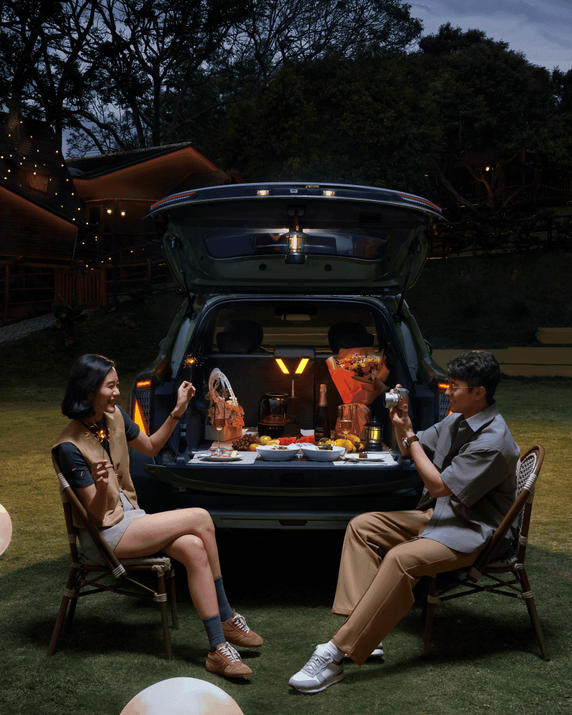A couple enjoying a picnic from the back of a car at night, seated on chairs with a table set up with food and drinks, surrounded by soft lighting in an outdoor setting.