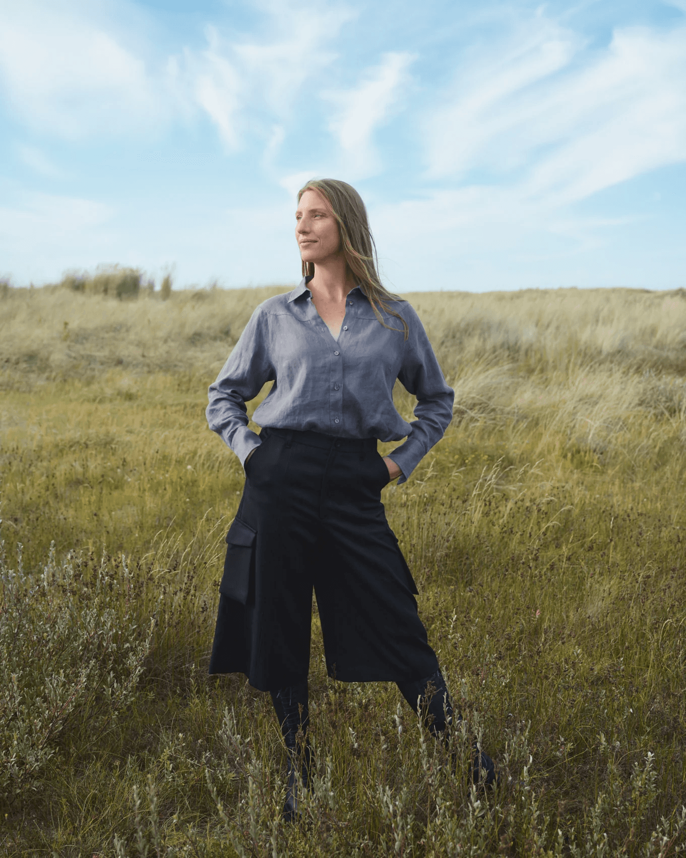 A person stands confidently in a grassy field under a bright blue sky with scattered clouds, wearing a blue shirt and black culottes, embodying a sense of freedom and nature.