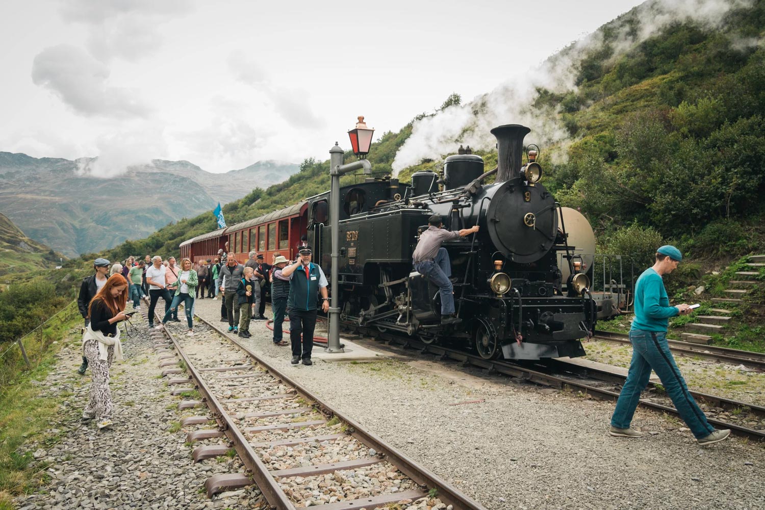 Das Foto zeigt eine historische, schwarze Dampflokomotive in schräger Frontansicht, die auf einem Schottergleis steht und zwei große, weiße Dampfwolken in die Luft stößt. Die Lokomotive verfügt über glänzende, messingfarbene Lampen und rote Details am vorderen Pufferblock, auf dem die weiße Beschriftung „DFB 704“ zu lesen ist. Rechts im Bild sind Ansätze von roten Personenwaggons zu erkennen, die an die Lok angehängt sind. Im Hintergrund erstreckt sich eine steile, grüne Berglandschaft mit Nadelbäumen und einem kleinen, alleinstehenden Holzhaus unter einem wolkenverhangenen Himmel.