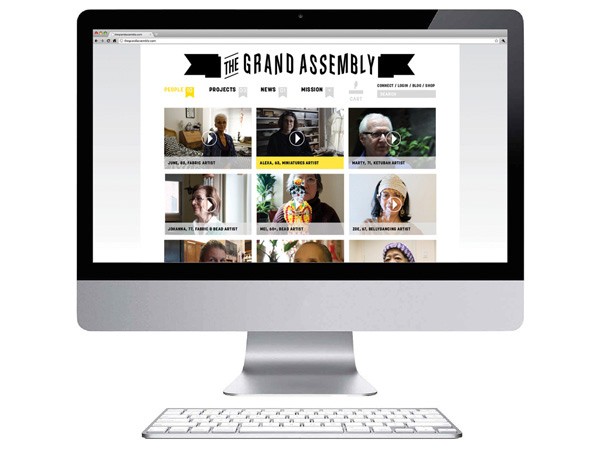 A silver Apple iMac computer displays a website titled “THE GRAND ASSEMBLY” on its screen. The website features eight thumbnail images of people, arranged in two rows of four, each labeled with a name and a small icon. The navigation bar includes tabs labeled “PEOPLE,” “PROJECTS,” “NEWS,” “MISSION,” “CONTACT,” and “SEARCH.” A white Apple keyboard is positioned below the computer.