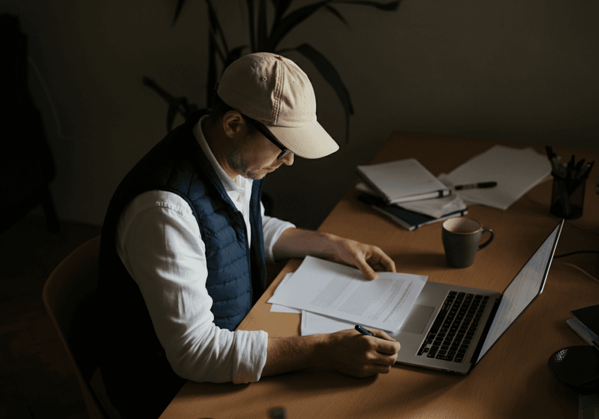 Man in a cap and glasses working at a wooden desk with documents and a laptop. The dimly lit room has a focused, thoughtful atmosphere.