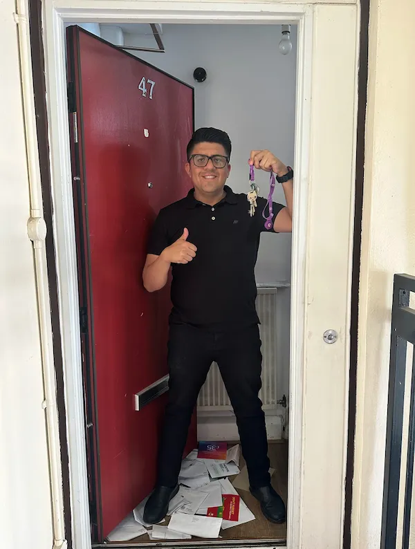 Man giving thumbs up while standing in a hallway next to a red door, wearing dark clothing