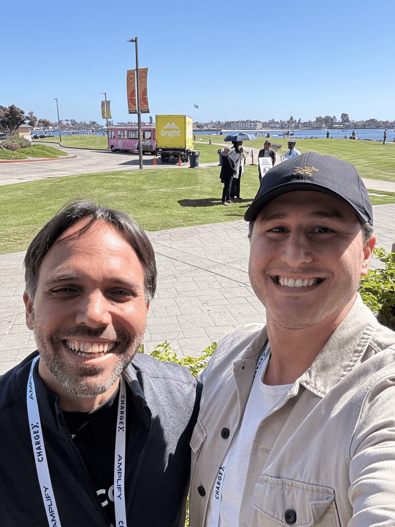 Two smiling men pose for a selfie outdoors with a scenic view in the background.