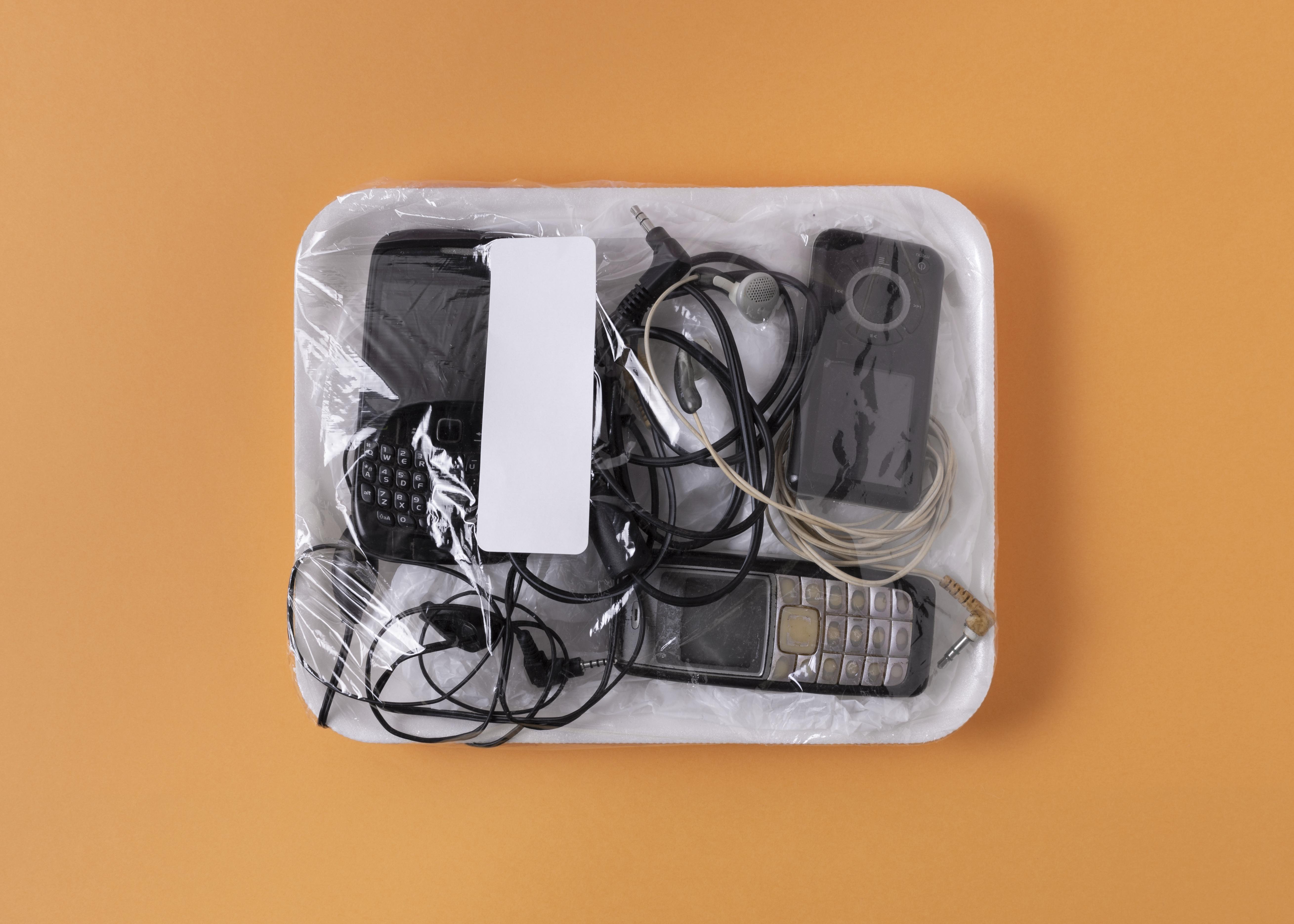 A clear plastic-wrapped tray containing old cell phones, headphones, and various cables on an orange background conveys a sense of technology storage.