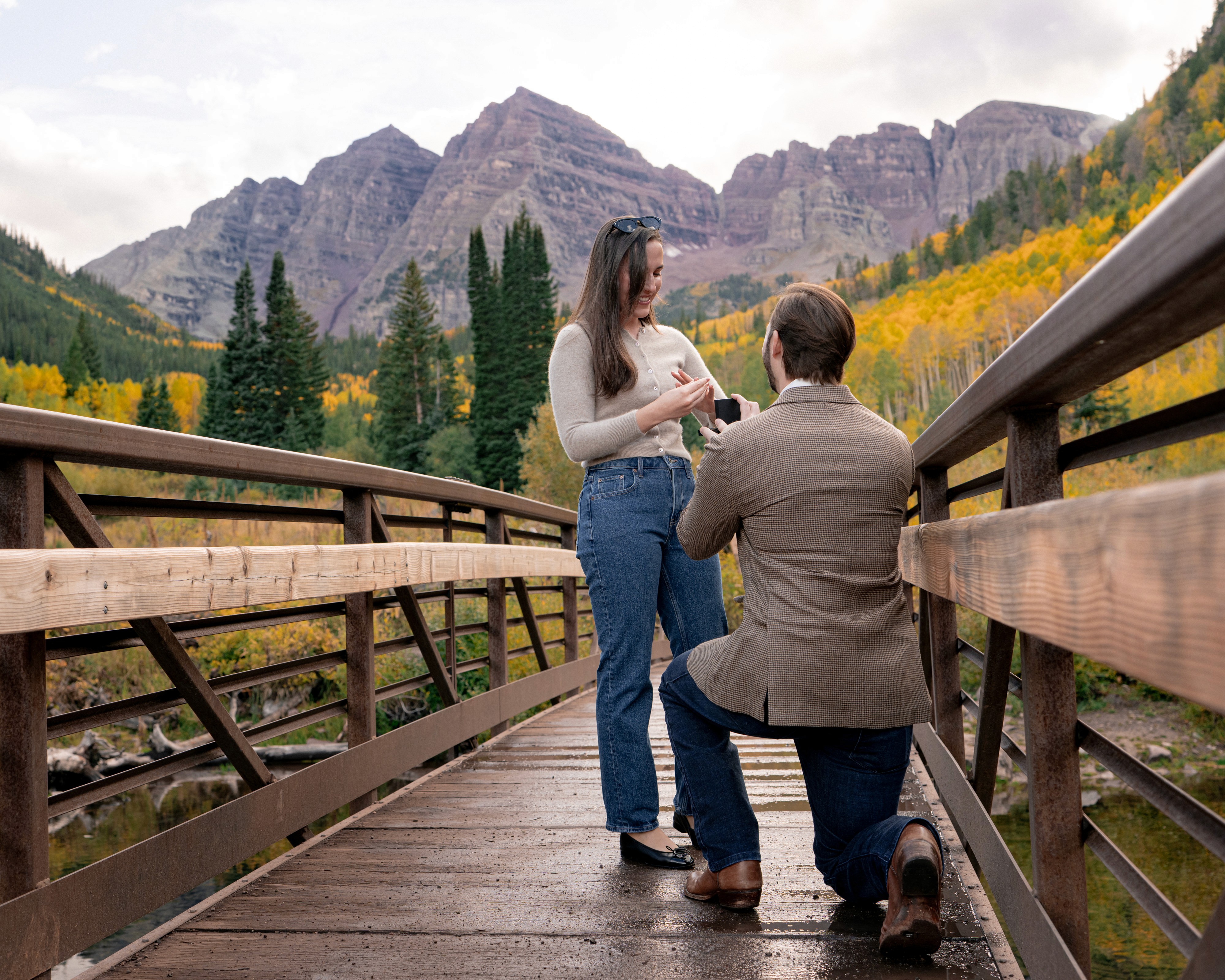 Tim kneels to propose to Lexi on a wooden bridge, with golden aspens and the Maroon Bells behind them.
