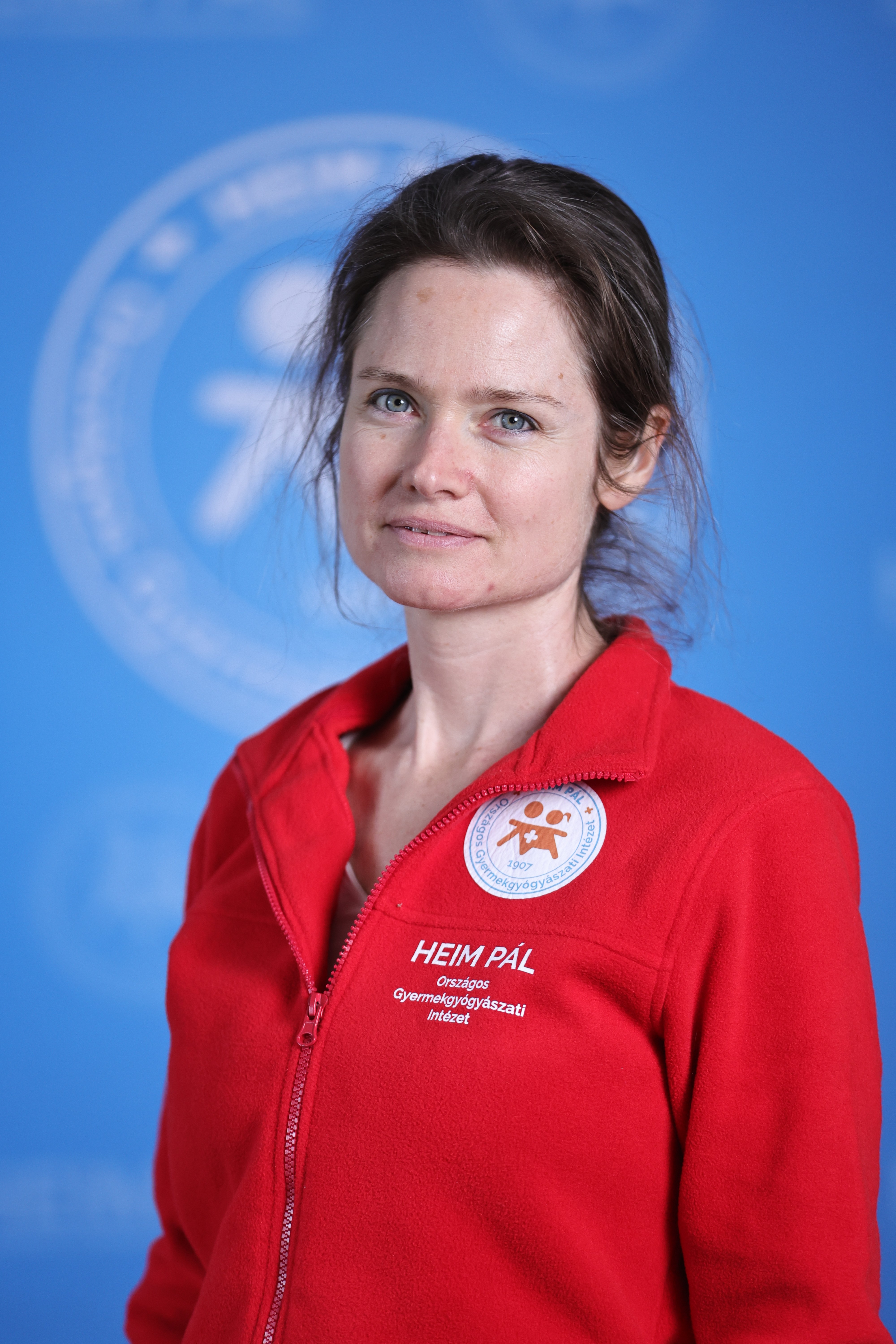 Hosszú, barna hajú nő piros Heim Pál feliratú kabátban áll kék háttér előtt.