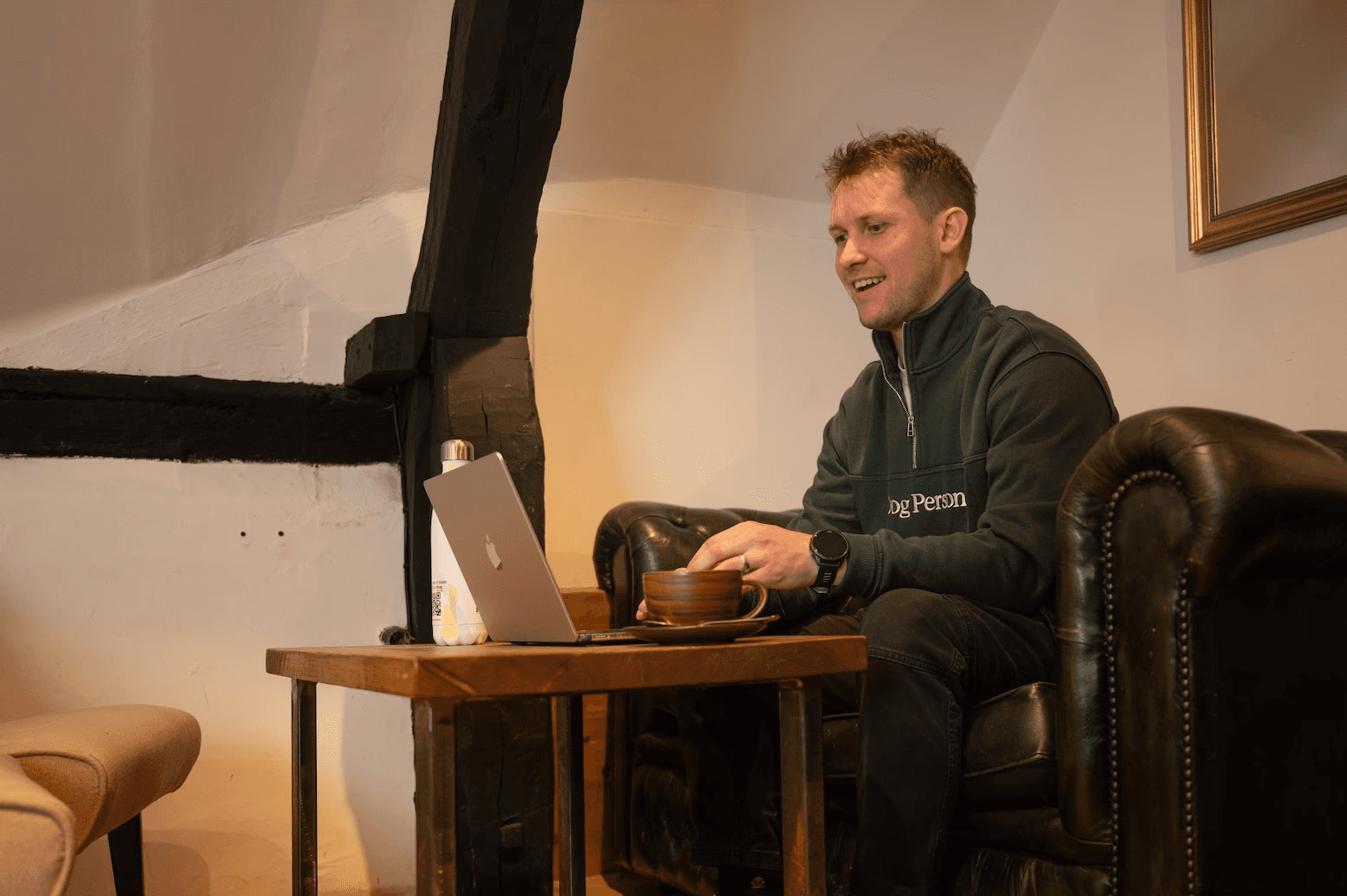 Dog Person sat on a chesterfield sofa, using their laptop. Beside the laptop is a coffee and a branded water bottle