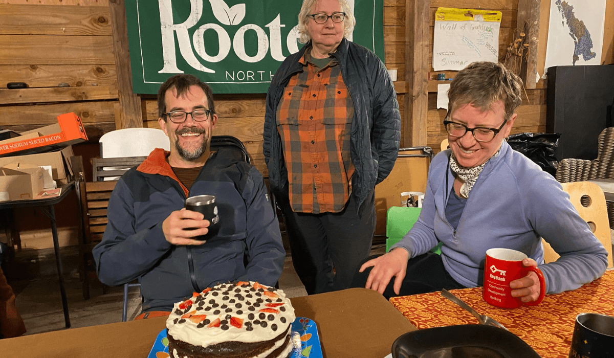 Three community members gather indoors around a homemade cake during a shared meal at the Rooted Northwest barn.