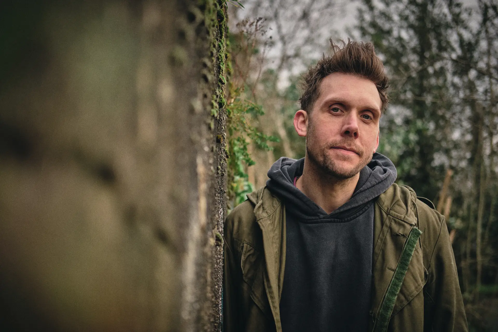 A young man stands thoughtfully against a blurred natural background, wearing a hoodie and a jacket.