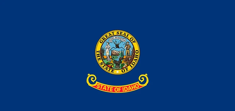 Flag of Idaho featuring a blue background with the state seal at the center. The seal depicts a miner, a woman, and symbols of agriculture and nature. Below, a red and yellow banner reads "State of Idaho."