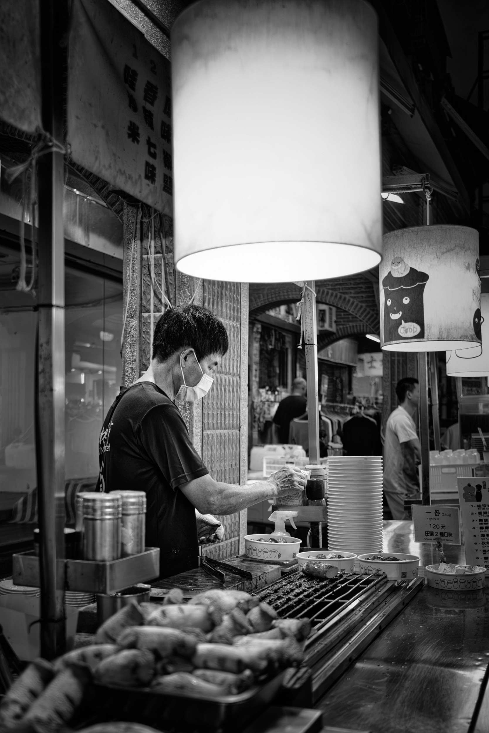Black and white photograph of a cook preparing bowls of food beneath a large hanging lamp at a street food stall.