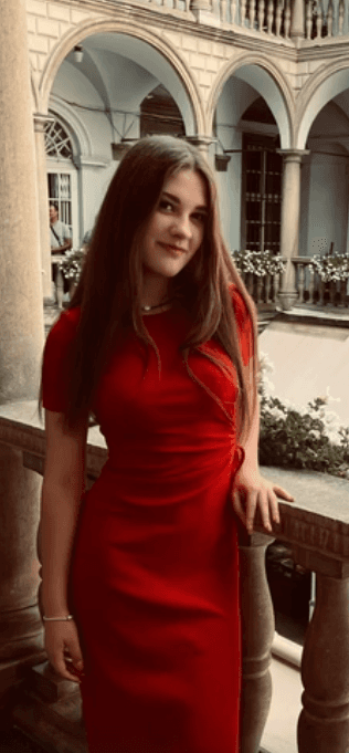 A woman in a red dress smiles while standing near an ornate balcony. Historic architecture is visible in the background.