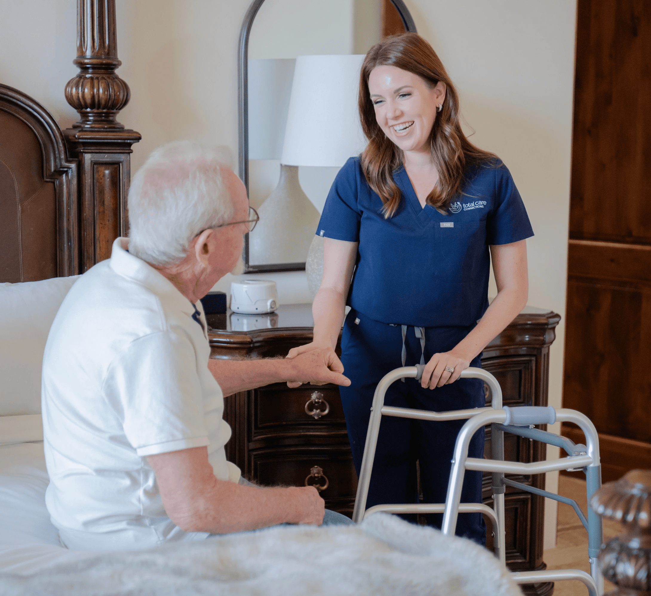 A woman stands beside a bed holding a man’s hand, a walker beside her.