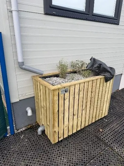 Wooden planter box against a wall, with a pipe nearby and some plants inside the box. Ground is paved.