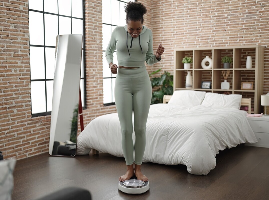 woman smiling as she stands on a scale in her room to check her results after working hard to lose weight on treadmill